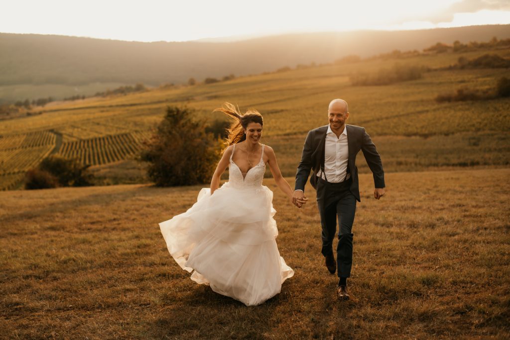 Morgane et Roberto, mariés souriants, courant dans un paysage de campagne, photo de mariage par Pauline Kupper, photographe Alsace